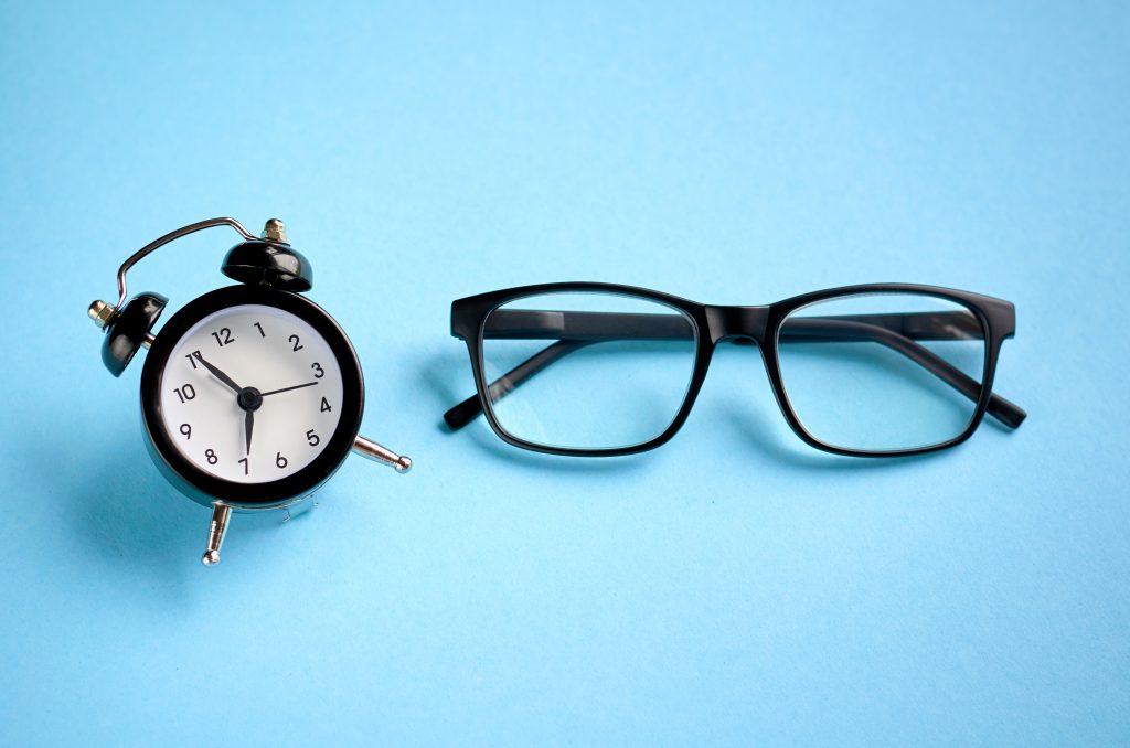 Black glasses and alarm clock on blue background composition.